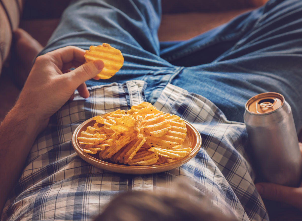 Bored guy lying on sofa eats crisps and drinks beer