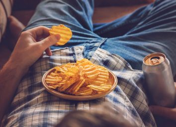 Bored guy lying down on couch eating potato chips and drinking a beer