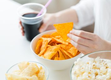 Woman reaching for chip and holding soda in processed junk food array on table with popcorn