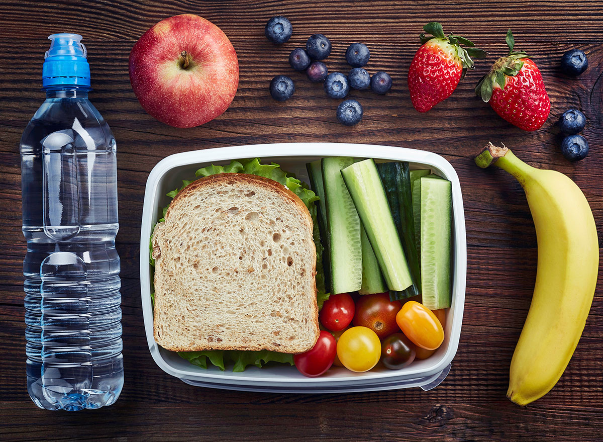sandwich in a container with cucumber sticks heirloom tomatoes surrounded by blueberries strawberries an apple a banana and a plastic water bottle