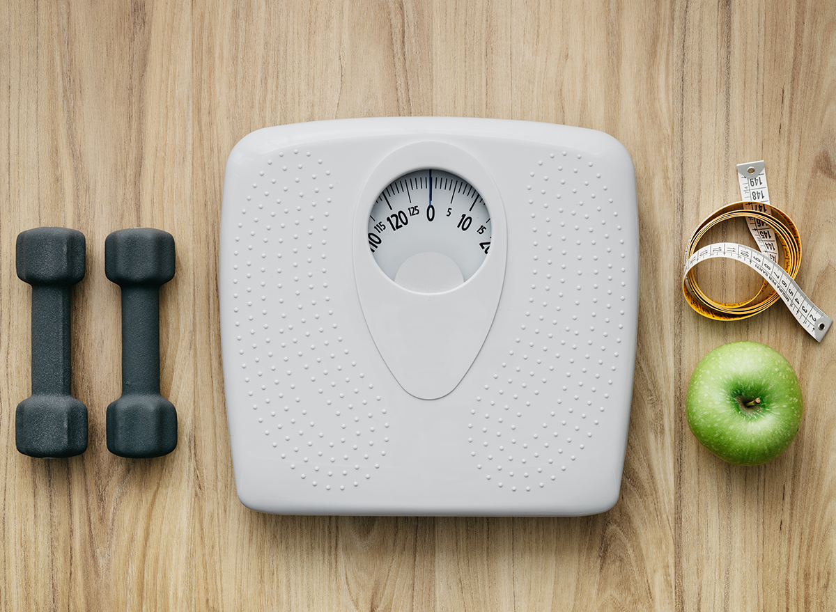 dumbbells sitting next to a scale with measuring tape and a green apple