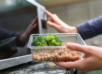 person putting container of broccoli and grains into the microwave