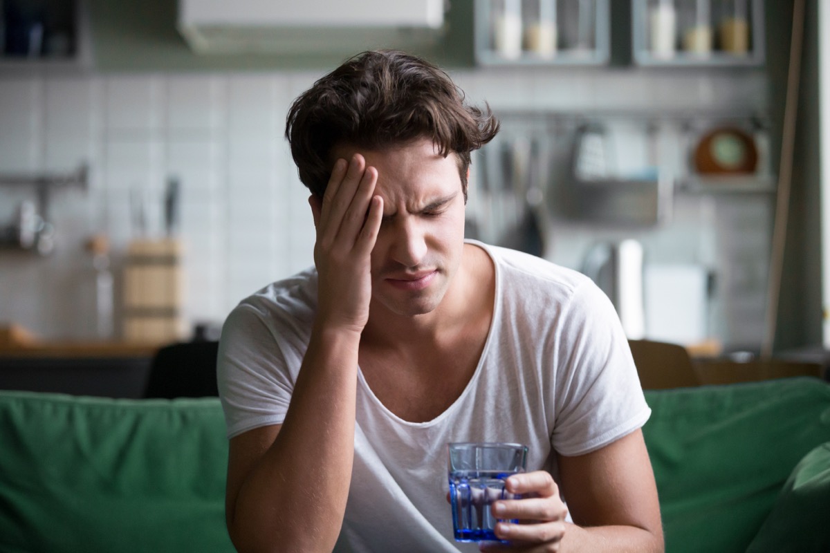 Young man suffering from strong headache or migraine sitting with glass of water in the kitchen, millennial guy feeling intoxication and pain touching aching head