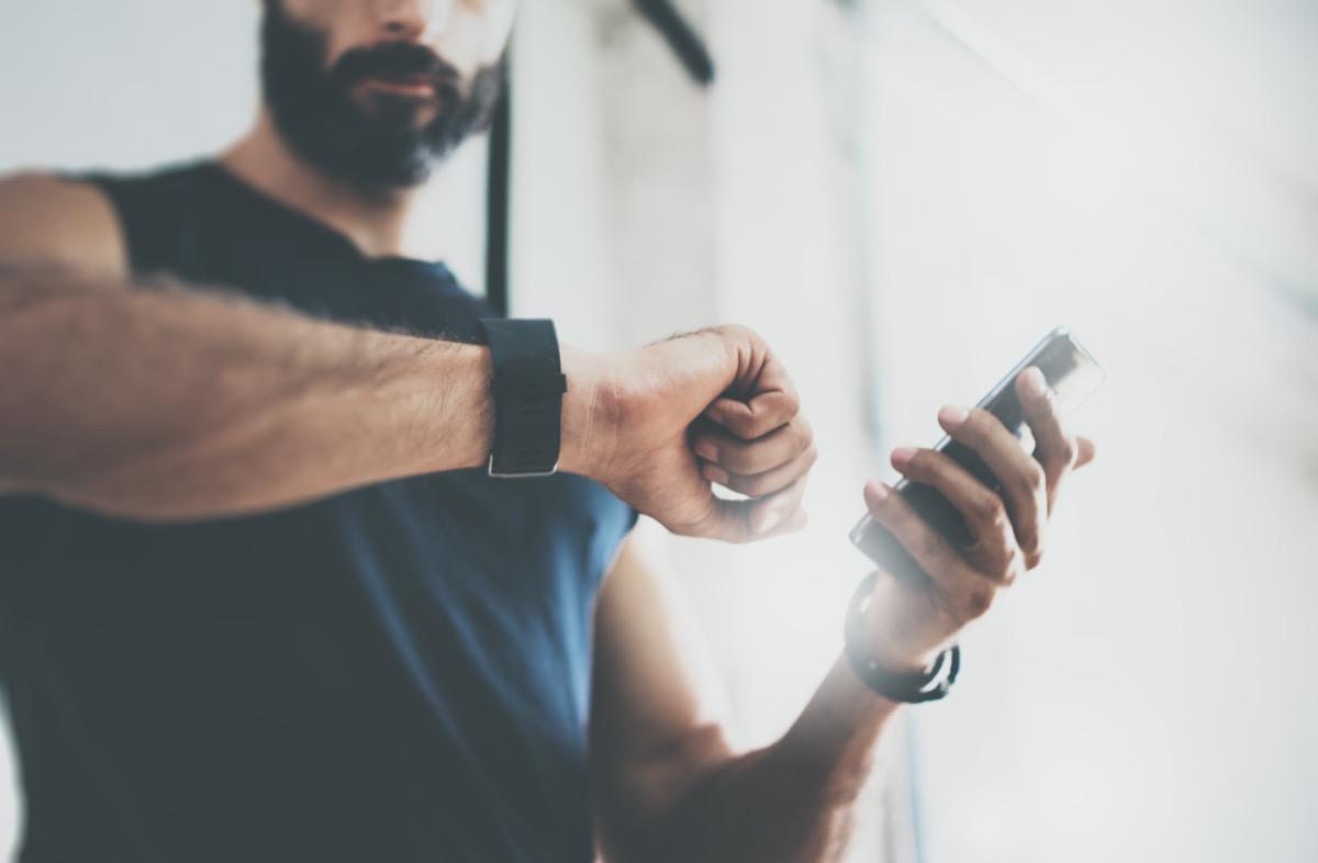 Close-up Shot Bearded Sportive Man After Workout Session Checks Fitness Results Smartphone.Adult Guy Wearing Sport Tracker Wristband Arm.Training hard inside gym.Horizontal bar background.Blurred - Image