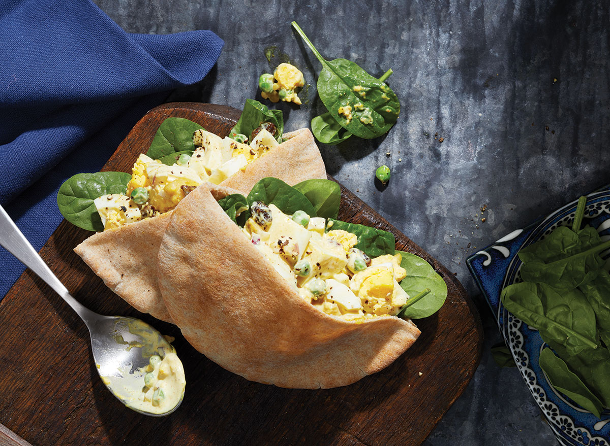curried egg salad in pita pocket with spoon and bowl of spinach on wooden serving board with blue linen napkin