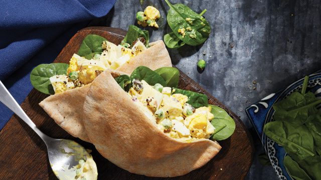 curried egg salad in pita pocket with spoon and bowl of spinach on wooden serving board with blue linen napkin