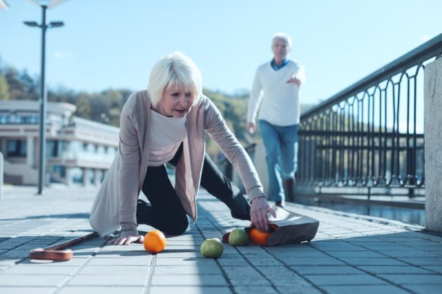 Senior lady standing on her knee and trying to pick up her groceries after falling down while her worried husband running to her in the background