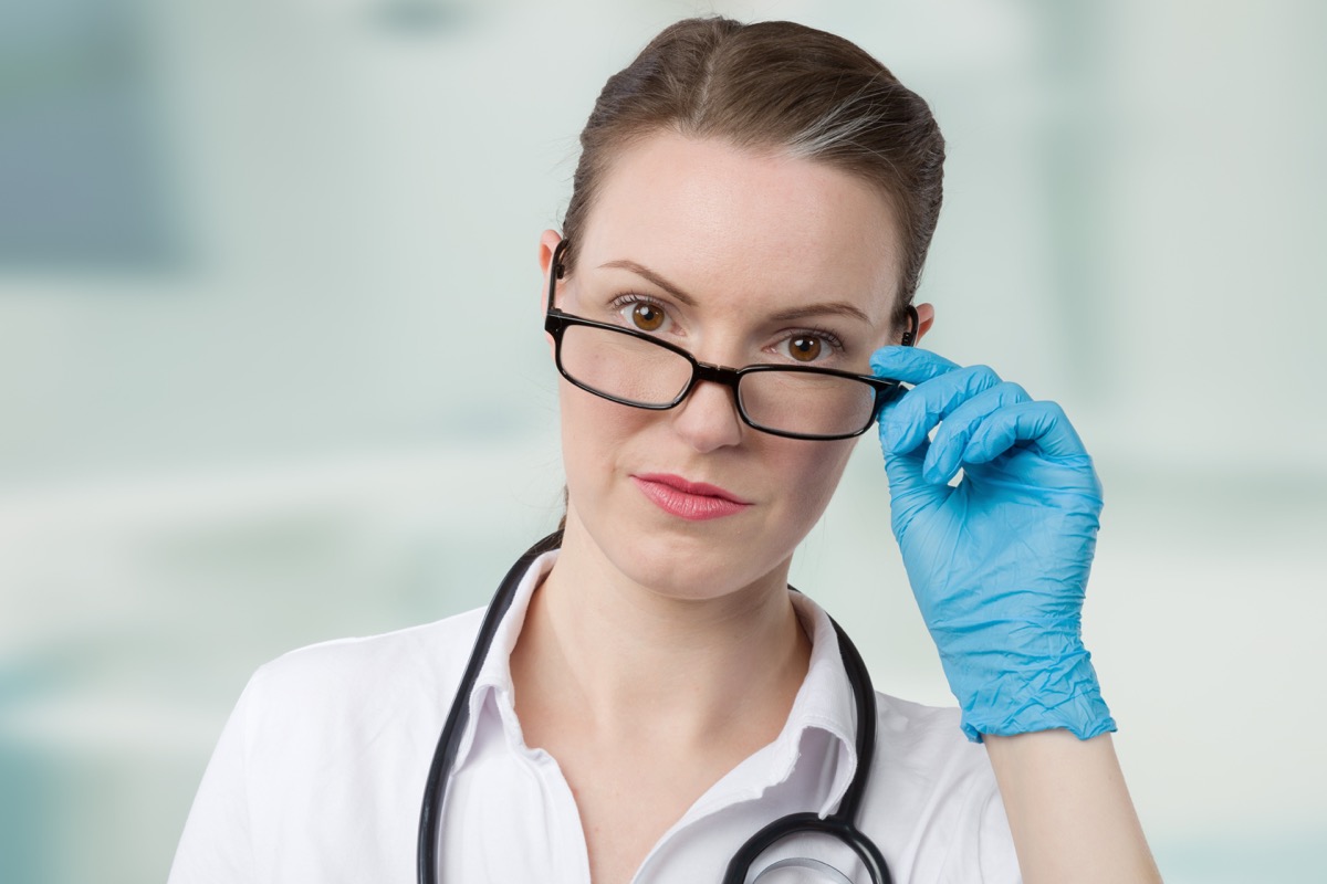 female doctor with blue medical gloves and a stethoscope looks over her glasses in front of a clinic room