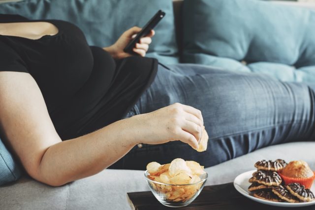 Obese woman lying on sofa with smartphone eating potato chips