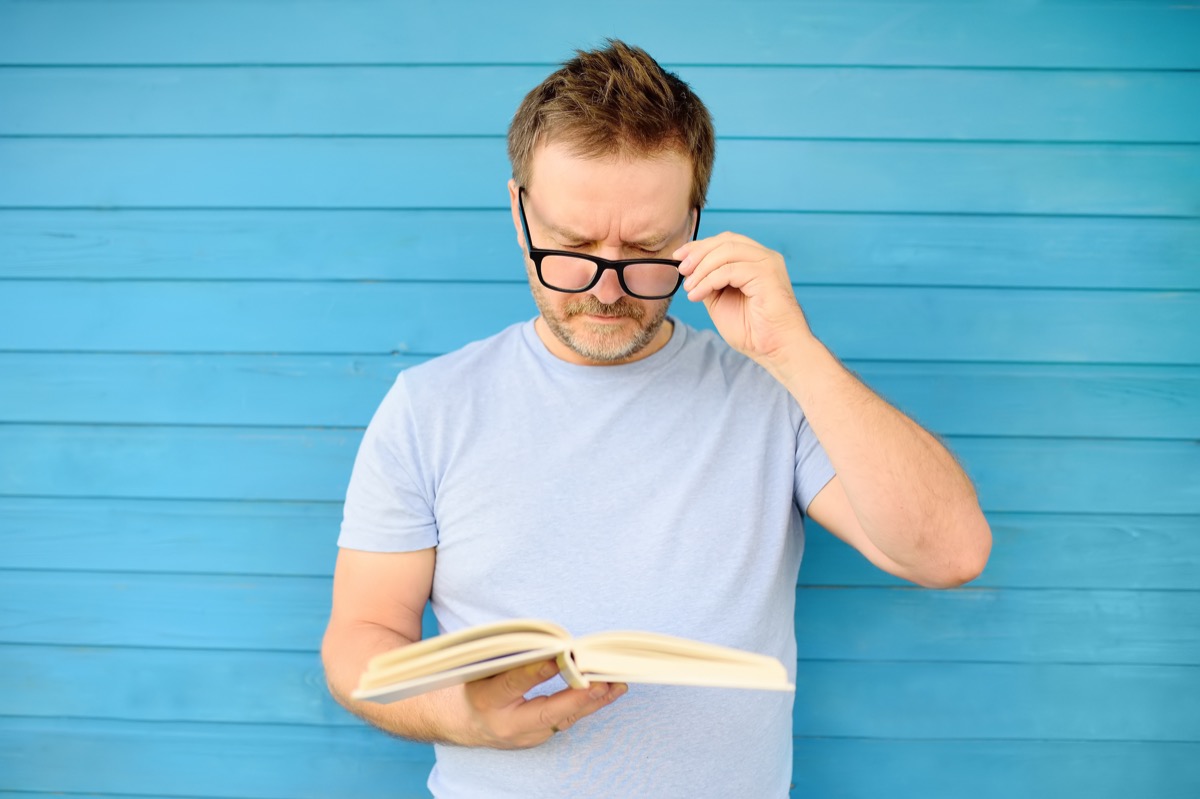 Portrait of mature man with big black eye glasses trying to read book but having difficulties seeing text because of vision problems. Problems disorder vision. - Image