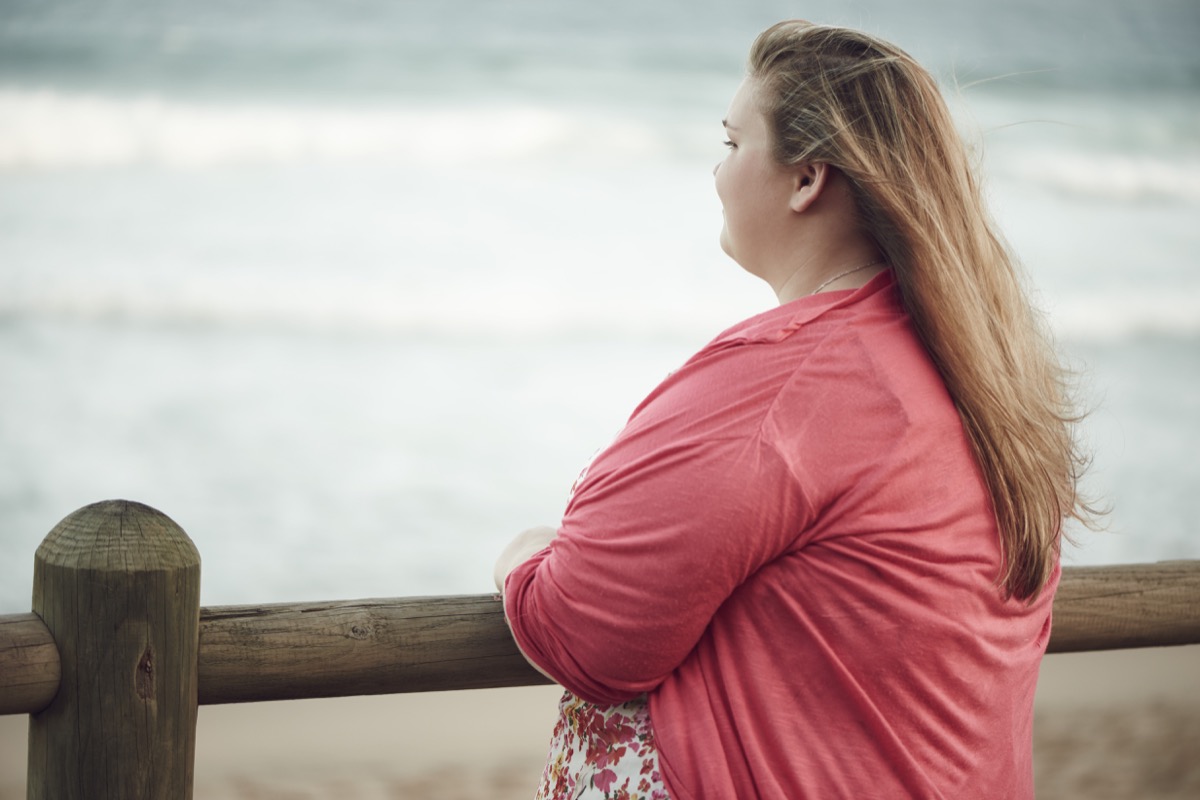 Young overweight caucasian adventurous teenage girl with blonde hair looking out at the ocean, while leaning on the wooden railing along the beach