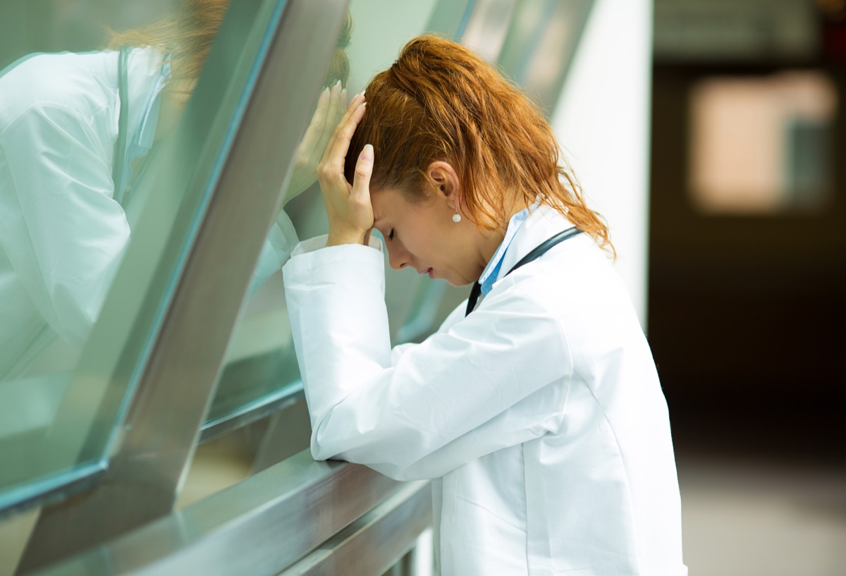 sad health care professional with headache, stressed, holding head against window glass. Nurse doctor with migraine overworked, overstressed isolated background hospital hall corridor