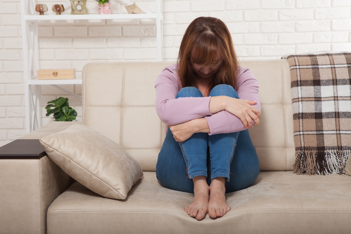 Sad middle aged woman sitting on sofa with clamped knees and crying