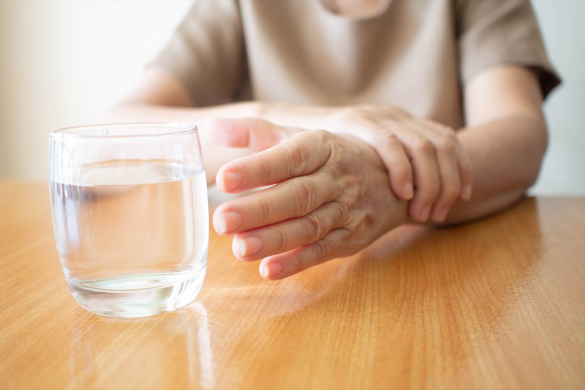 Elderly woman hands w/ tremor symptom reaching out for a glass of water on wood table. Cause of hands shaking include Parkinson's disease, stroke or brain injury. Mental health neurological disorder