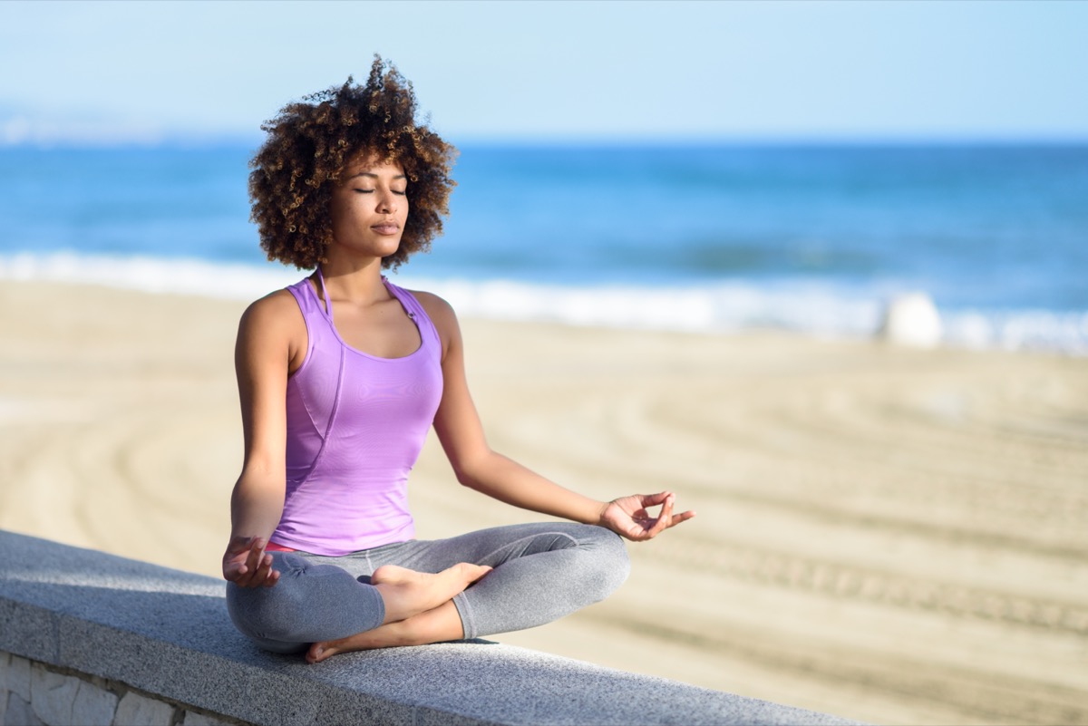 Black woman, afro hairstyle, doing yoga asana in the beach with eyes closed. Young Female wearing sport clothes in lotus pose with defocused background