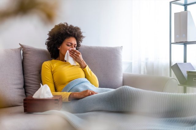 A woman sneezes in a handkerchief in the living room. A woman blowing her nose on the couch at home in the living room. An African-American woman with a handkerchief is sitting on the couch at home