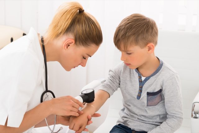 Female Doctor Examining Skin Of Little Boy With Dermatoscope