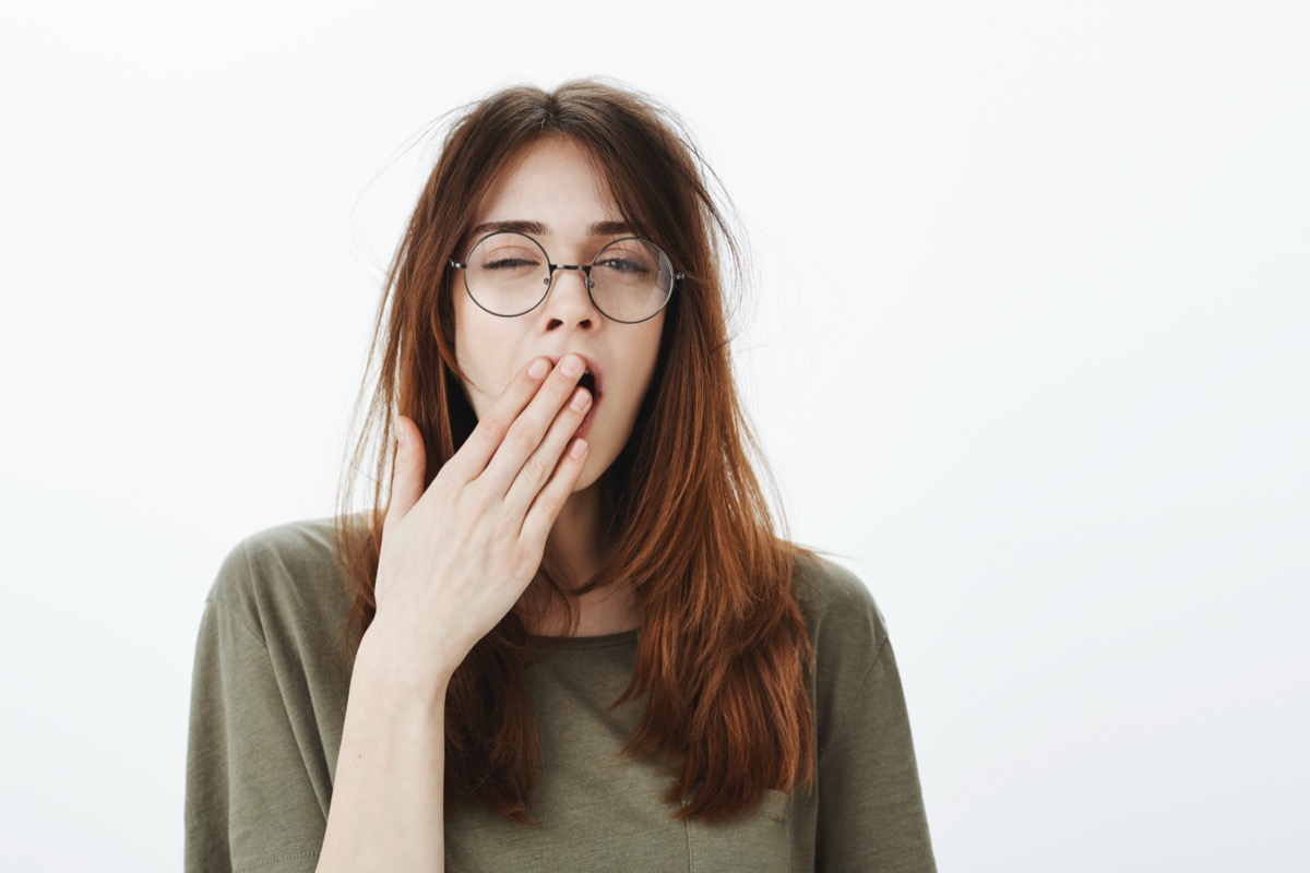 Shot of sleepy woman with messy brown hair, wearing glasses, feeling tired after night without sleep, yawning, covering opened mouth with palm