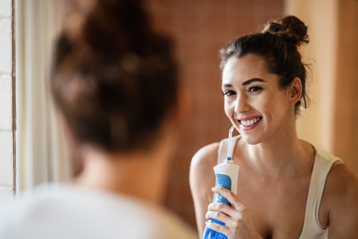 woman using dental water flosser while cleaning teeth and looking herself in bathroom mirror