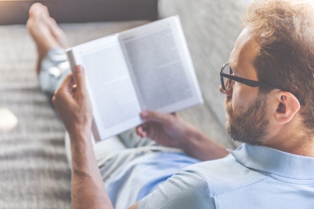 Mann in Freizeitkleidung und Brille, der ein Buch liest, während er zu Hause auf der Couch liegt
