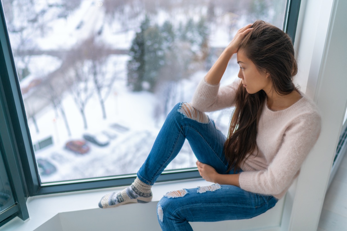 Winter depressed sad girl lonely by home window looking at cold weather upset unhappy