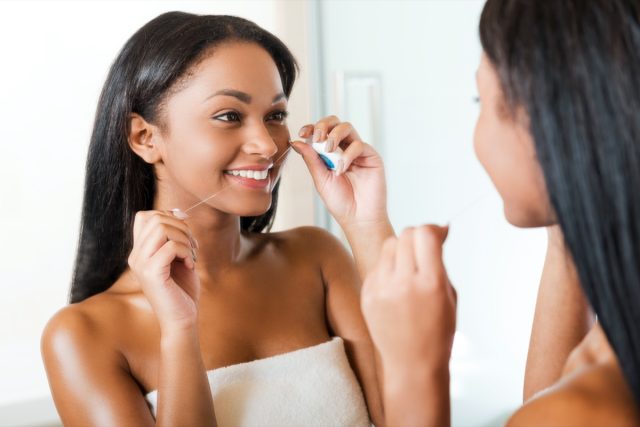 femeie care curăța dinții cu ața dentară și zâmbește în timp ce stă lângă oglinda din baie