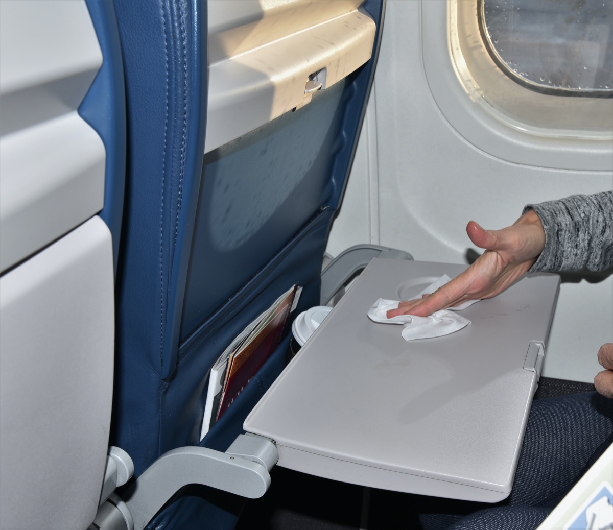 A woman sitting by a window seat is wiping down a germ laden dirty airplane tray with an antibacterial wipe