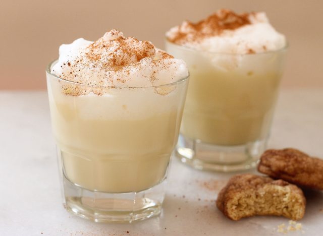 two cups of eggnog sprinkled with nutmeg next to two cookies
