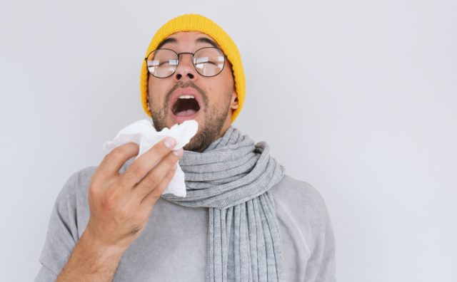 Ill man wearing grey sweater, yellow hat and spectacles, blowing nose and sneeze into tissue