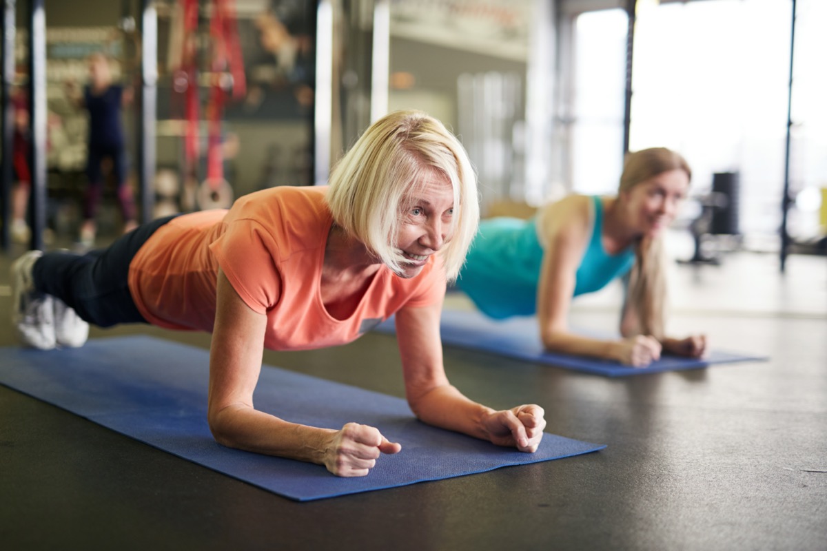 Mature blonde active woman standing in plank on mat during workout in contemporary fitness center.