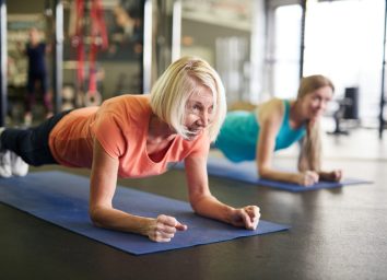 Mature blonde active woman standing in plank on mat during workout in contemporary fitness center.