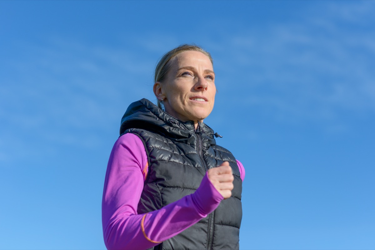 donna di mezza età, jogging in inverno in una low angle view contro un sole azzurro del cielo in un sano stile di vita attivo