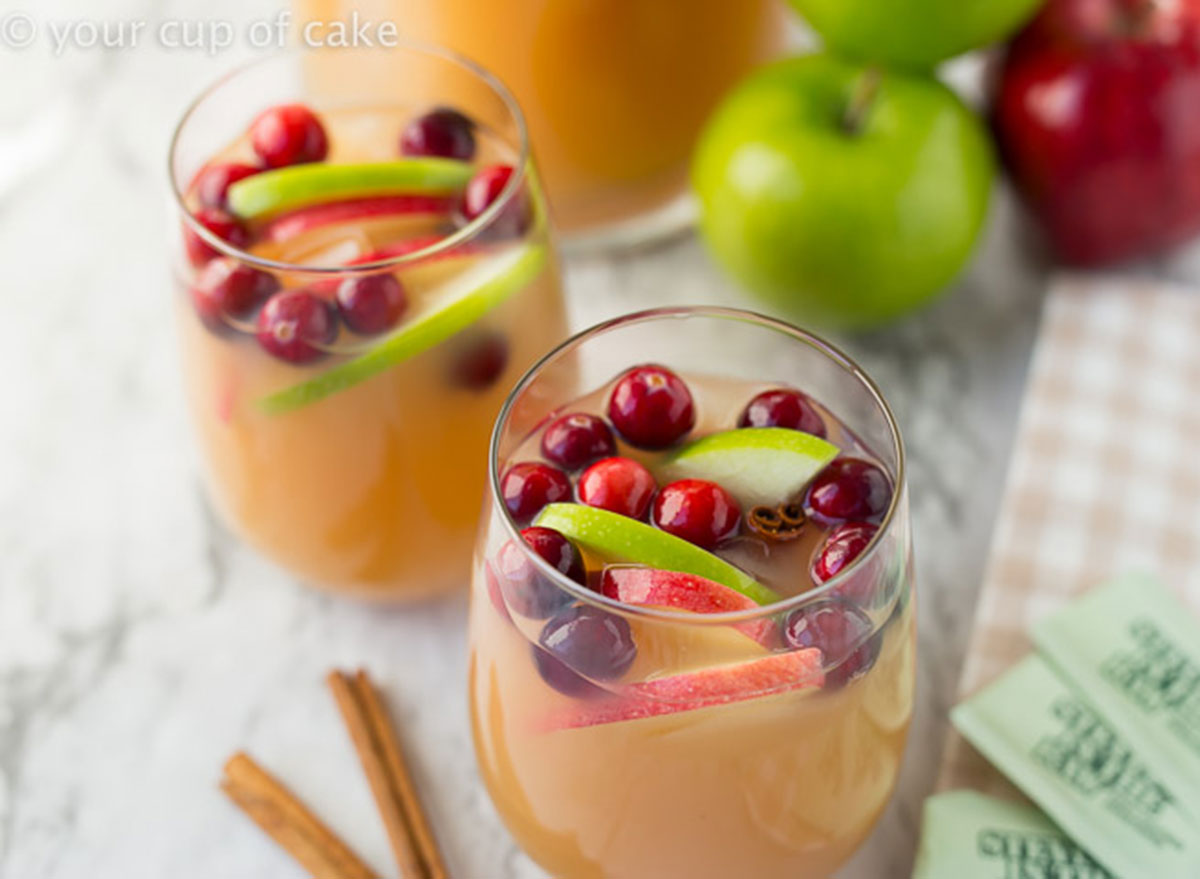 two glasses of sparkling caramel apple cider with cinnamon sticks next to apples on marble table