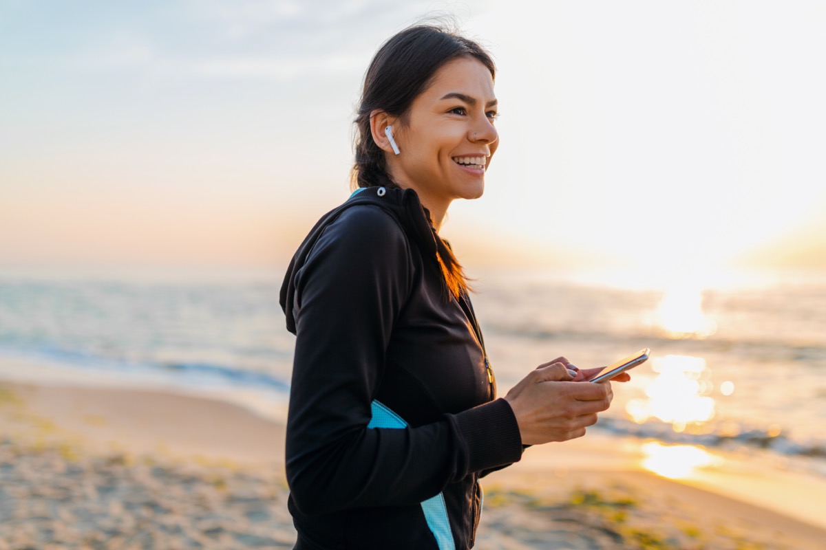 woman doing sport exercises on morning sunrise beach in sports wear, healthy lifestyle, listening to music on wireless earphones holding smartphone, smiling happy