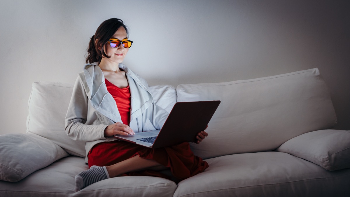 woman with blue light blocking glasses (yellow lenses) working with laptop on white sofa