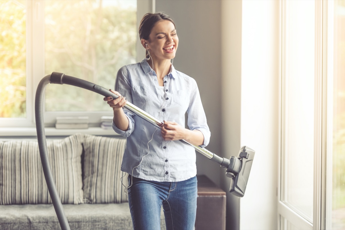 woman in earphones is imitating playing guitar using a vacuum cleaner and smiling while cleaning her house