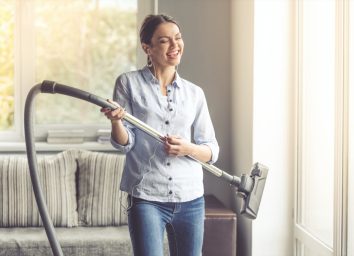 woman in earphones is imitating playing guitar using a vacuum cleaner and smiling while cleaning her house