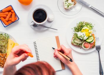 Woman writing in food journal with egg toast carrots coffee on table