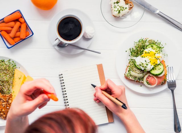 Woman writing in food diary with egg toast carrots coffee on table