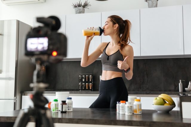 Satisfied healthy young girl recording her video blog episode about healthy food additives while standing at the kitchen at home and showing thumbs up