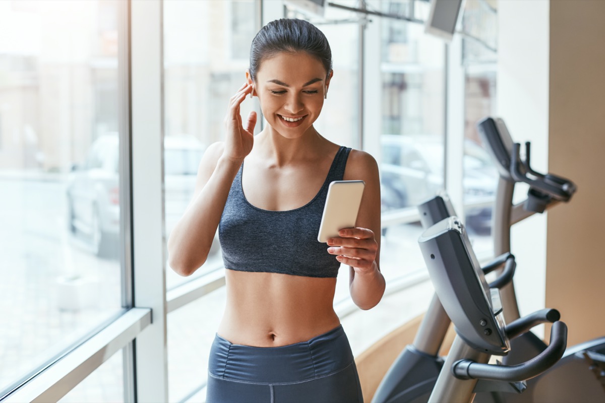 woman in sportswear choosing music for fitness on her smartphone while standing against treadmills at gym