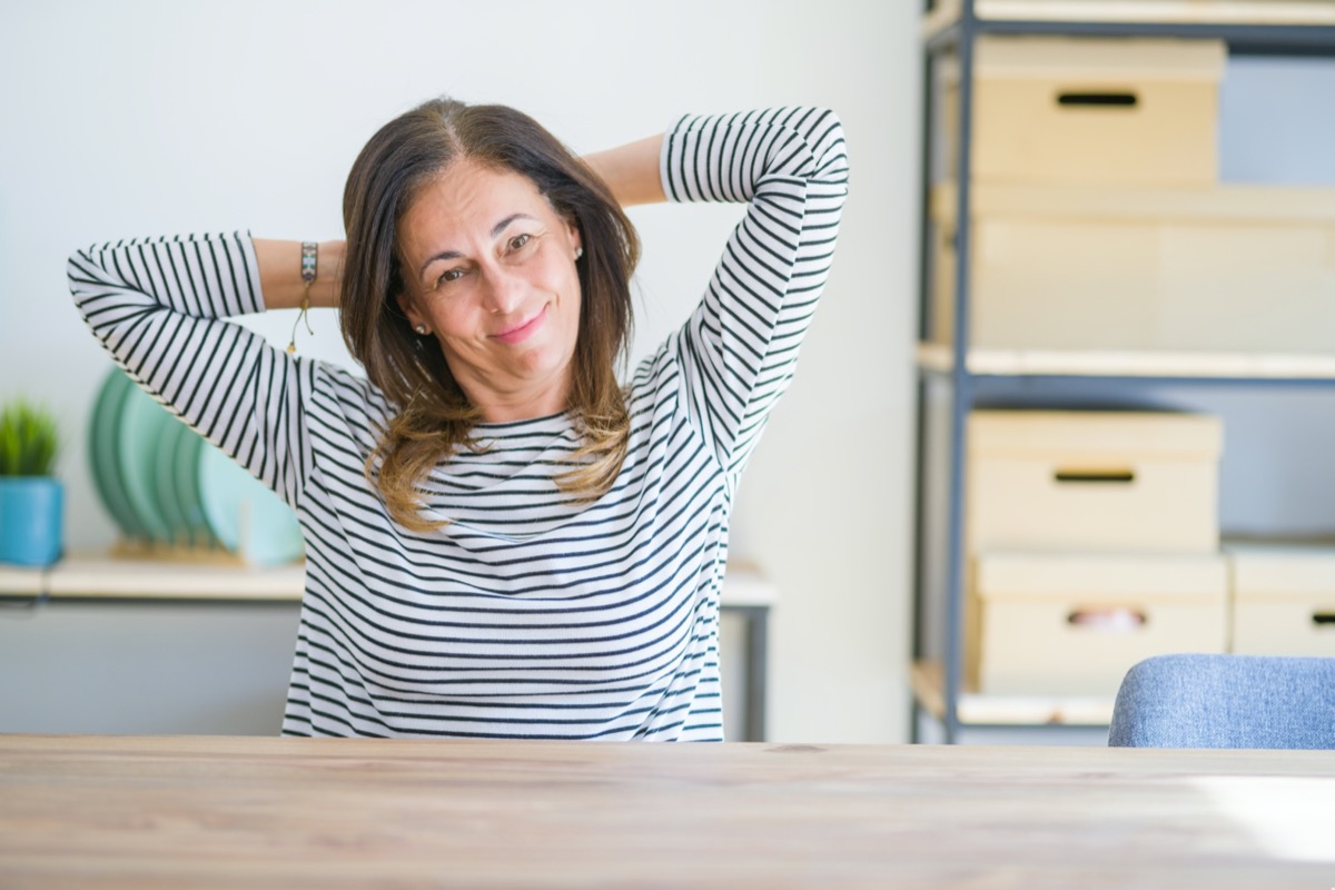 Middle age senior woman sitting at the table at home relaxing and stretching, arms and hands behind head and neck smiling happy