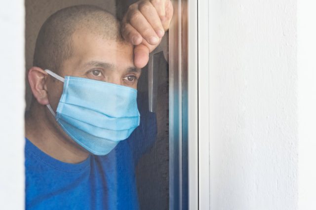 Sick man with face mask looking out the window being quarantined at home