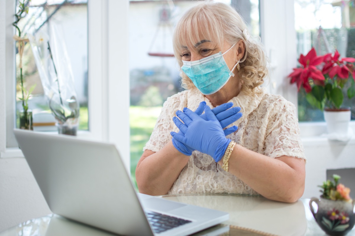 Scared mid age woman wearing protective mask and gloves reading news on laptop at home