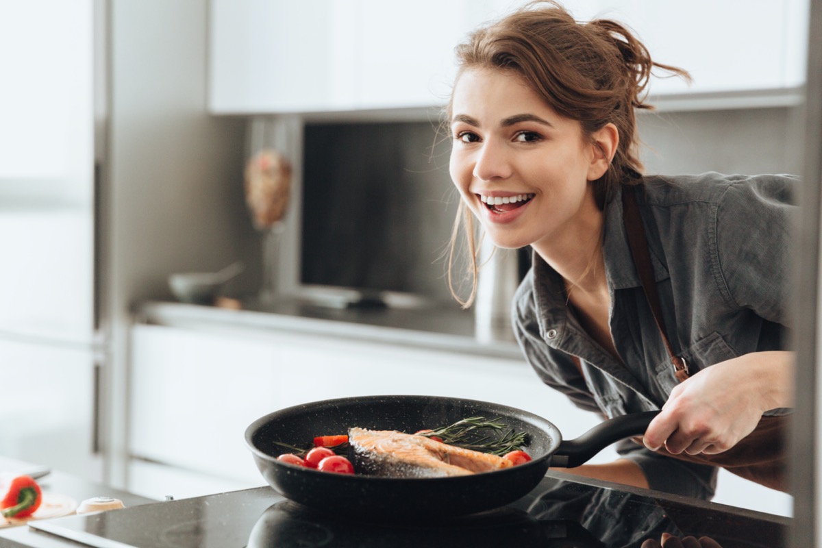 young happy lady standing in kitchen while cooking fish