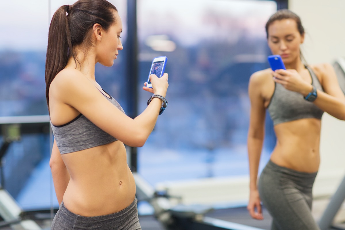 woman with smartphone taking mirror selfie in gym