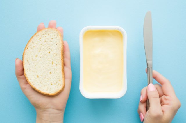 Woman's hands holding slice of white bread and knife. Opened plastic pack of light yellow margarine on pastel blue desk