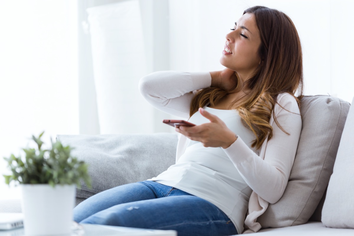 Shot of tired young woman with neck pain holding her mobile phone at home