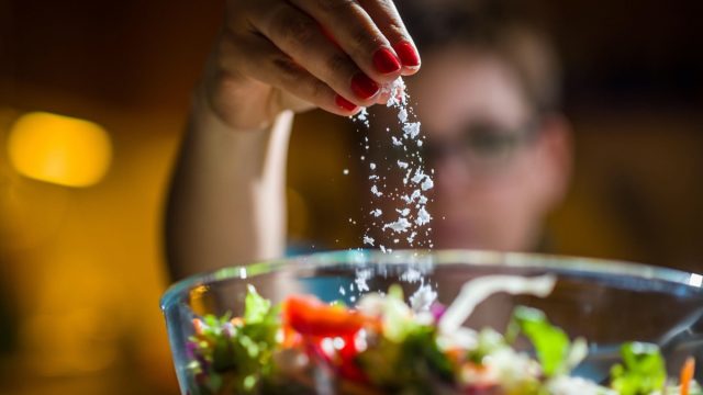 Woman preparing healthy salad in kitchen, adding salt to the bowl