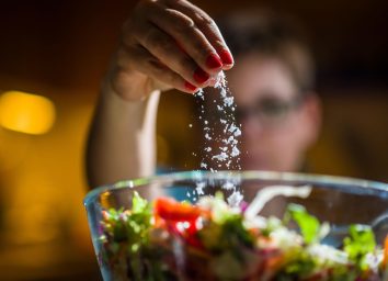 Woman preparing healthy salad in kitchen, adding salt to the bowl